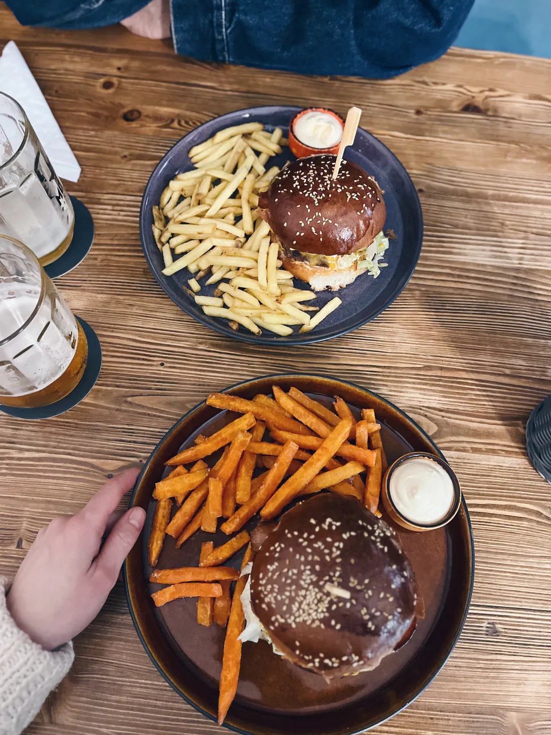 Dva burgery s klasickými i batátovými hranolky a sklenicemi piva na dřevěném stole