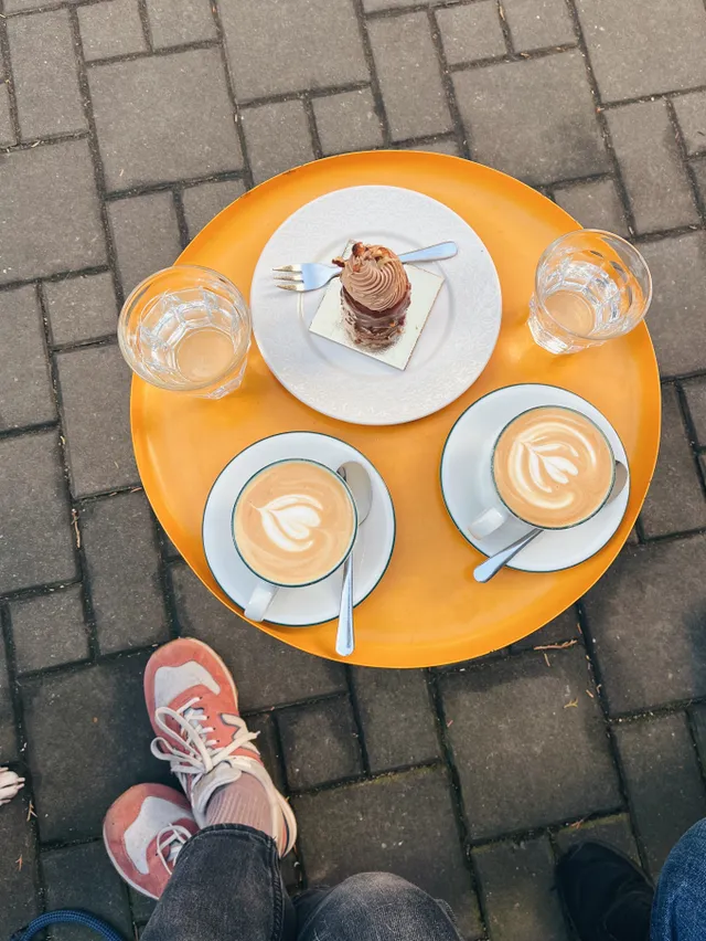 Oranžový stolík venku s dvěma cappucciny, vodou a čokoládovým dortíkem na talířku