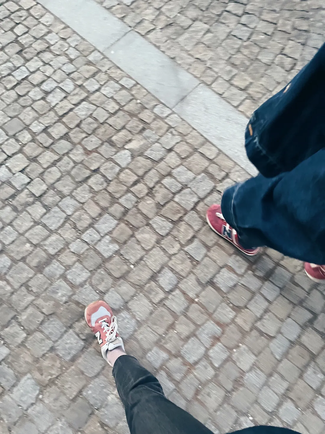 Nohy dvou lidí v červených teniskách New Balance kráčejících po dlážděné ulici