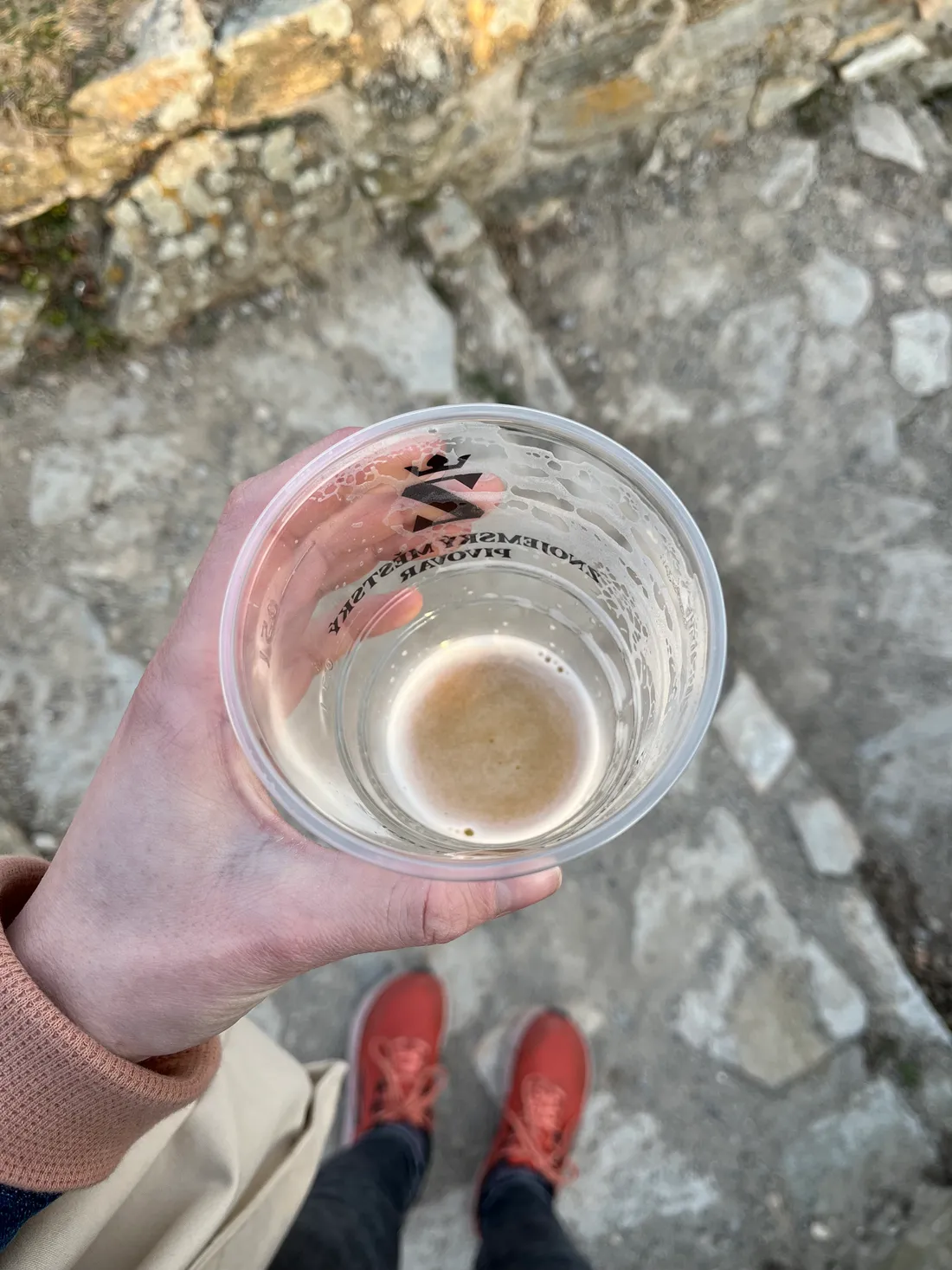 Ruka drží kelímek piva Znojemského městského pivovaru, v pozadí kamenná cesta
