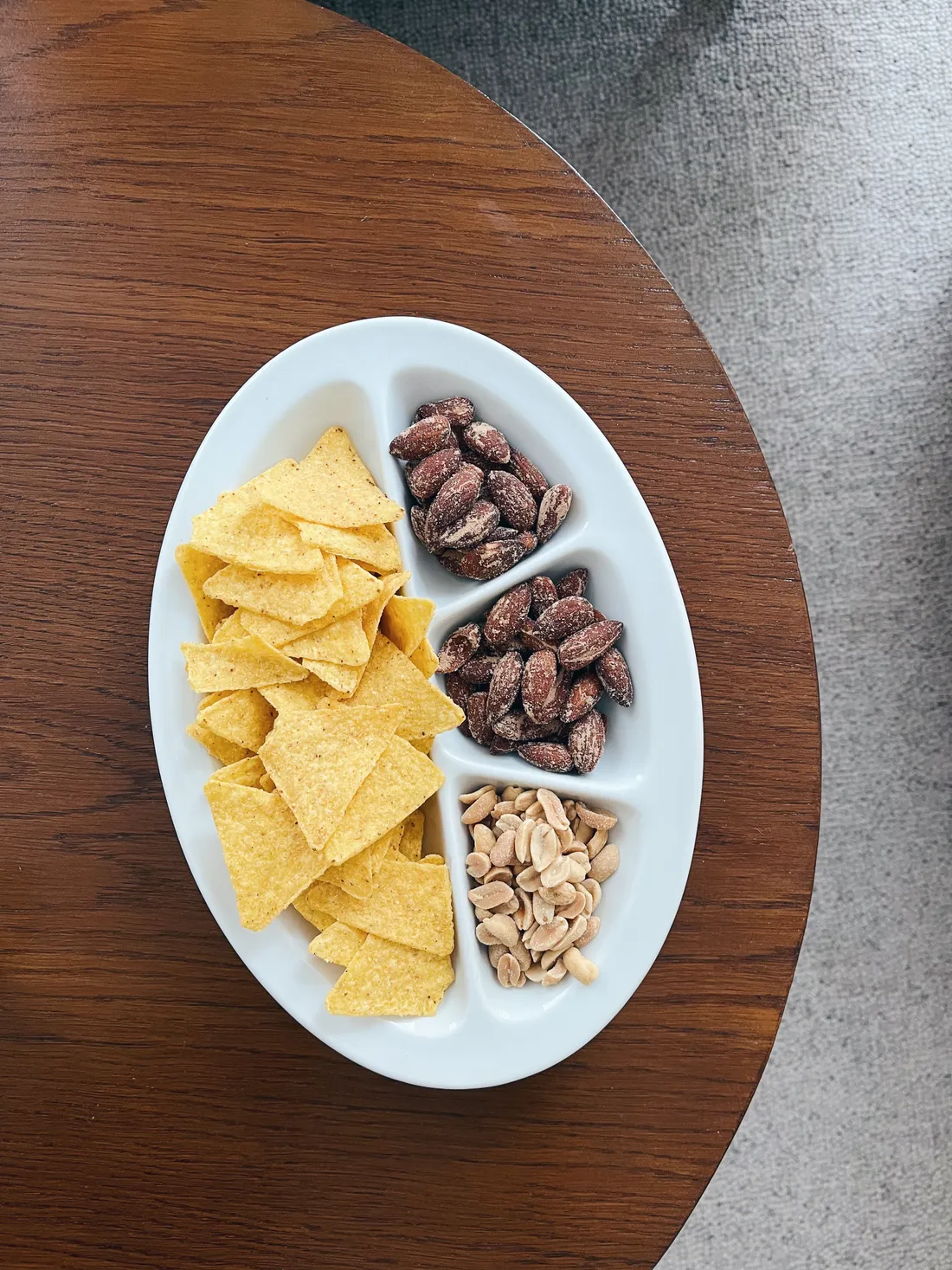Bílá miska se čtyřmi přihrádkami: nachos, mandle a arašídy na dřevěném stole