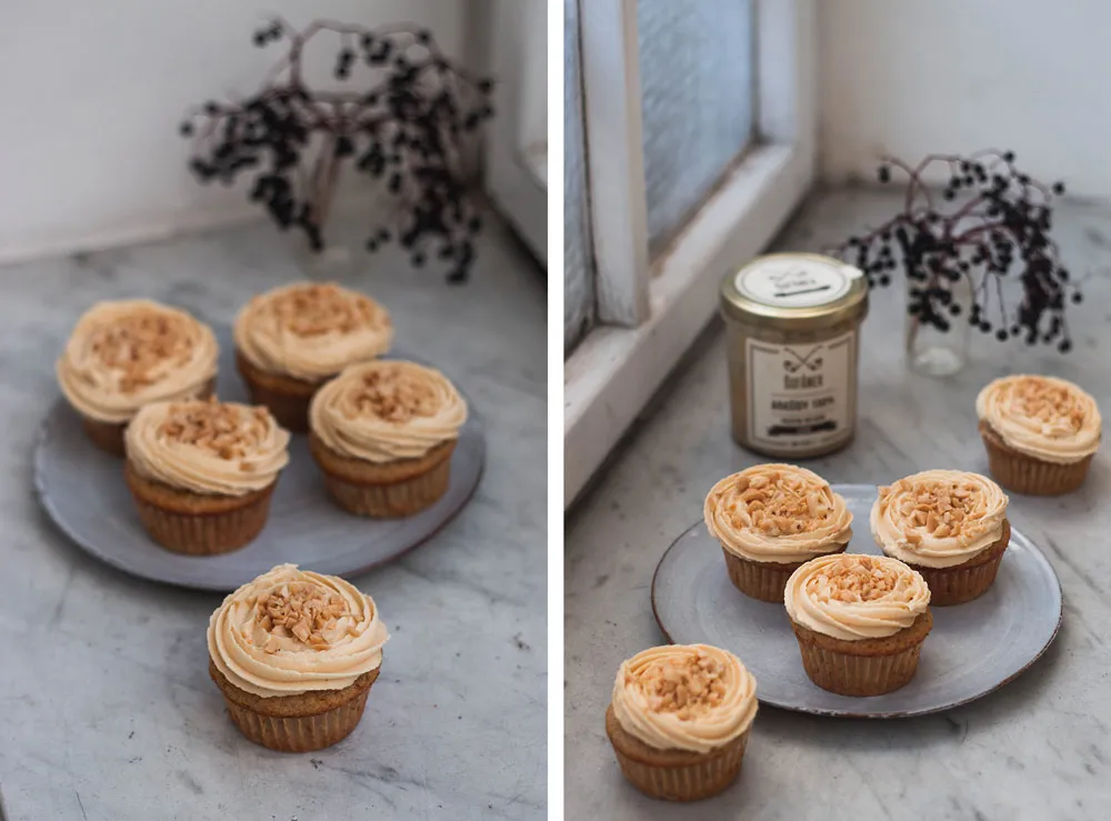 Banánové cupcakes s krémem z arašídového másla na mramorovém parapetu; v pozadí sklenice másla Šufan u světlého okna.