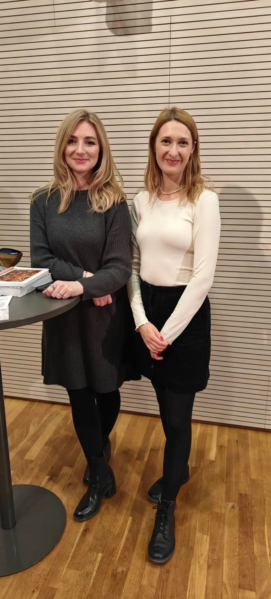 Dvě usmívající se ženy – blond v šedém úpletu a hnědovlasá v krémovém topu – u dřevěné stěny