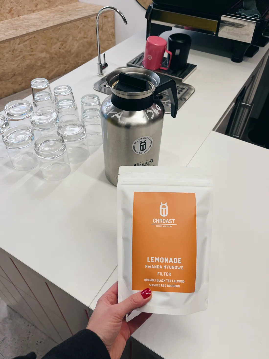 Ruka drží sáček kávy Chroast Lemonade z Rwandy u pultu s karafou a skleničkami v kavárně