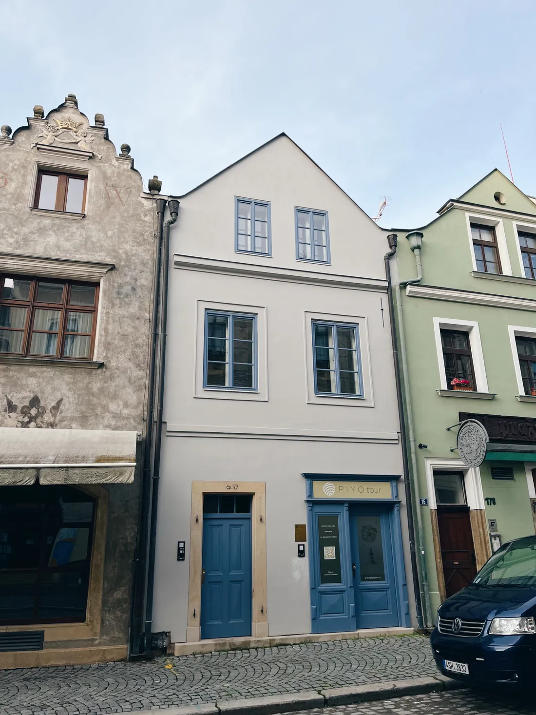 Řada historických domů na dlážděné ulici, uprostřed šedý dům s modrými okny a dveřmi