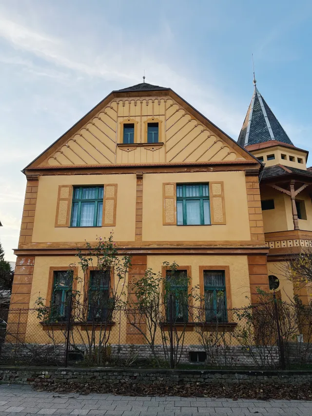 Žlutá historická vila se zelenými okny a okrasným štítem pod světle modrou oblohou