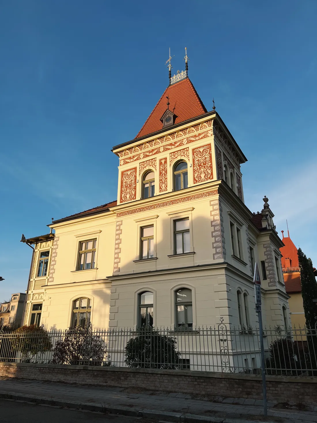 Velká historická vila s věží a červenou střechou ve večerním slunci za kovovým plotem