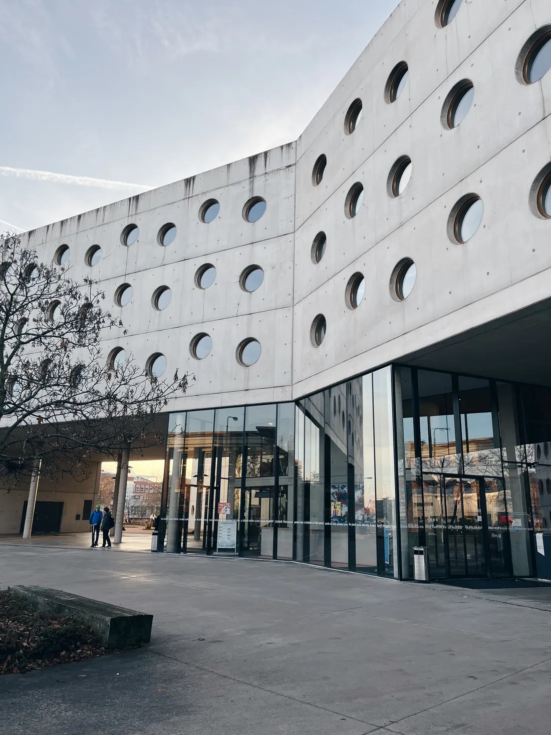 Betonová knihovna v Hradci Králové s mříží kulatých oken na dlážděném prostranství před vchodem