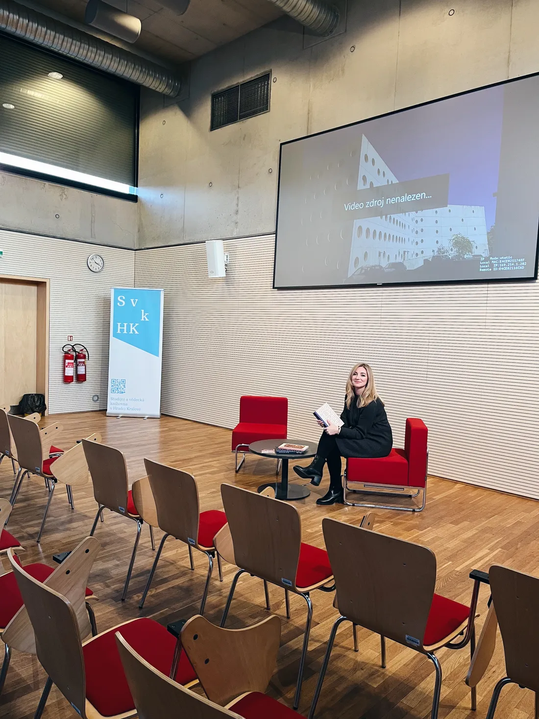 Blond žena v černých šatech sedí v červeném křesle před řadami židlí a projekční obrazovkou