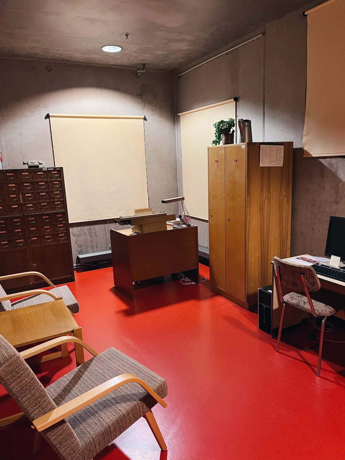 Retro pracovna s červenou podlahou, dřevěným nábytkem, psacím strojem a kartotékou u betonové zdi