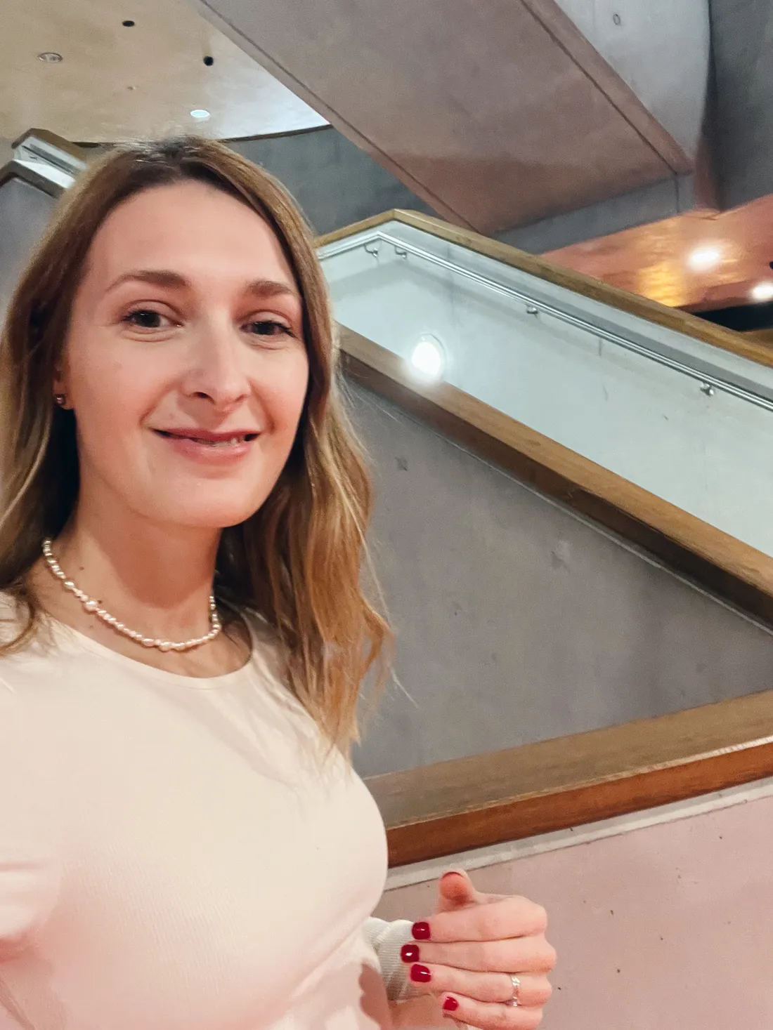 Selfie usmívající se ženy s perlovým náhrdelníkem u schodiště s betonovou stěnou v knihovně