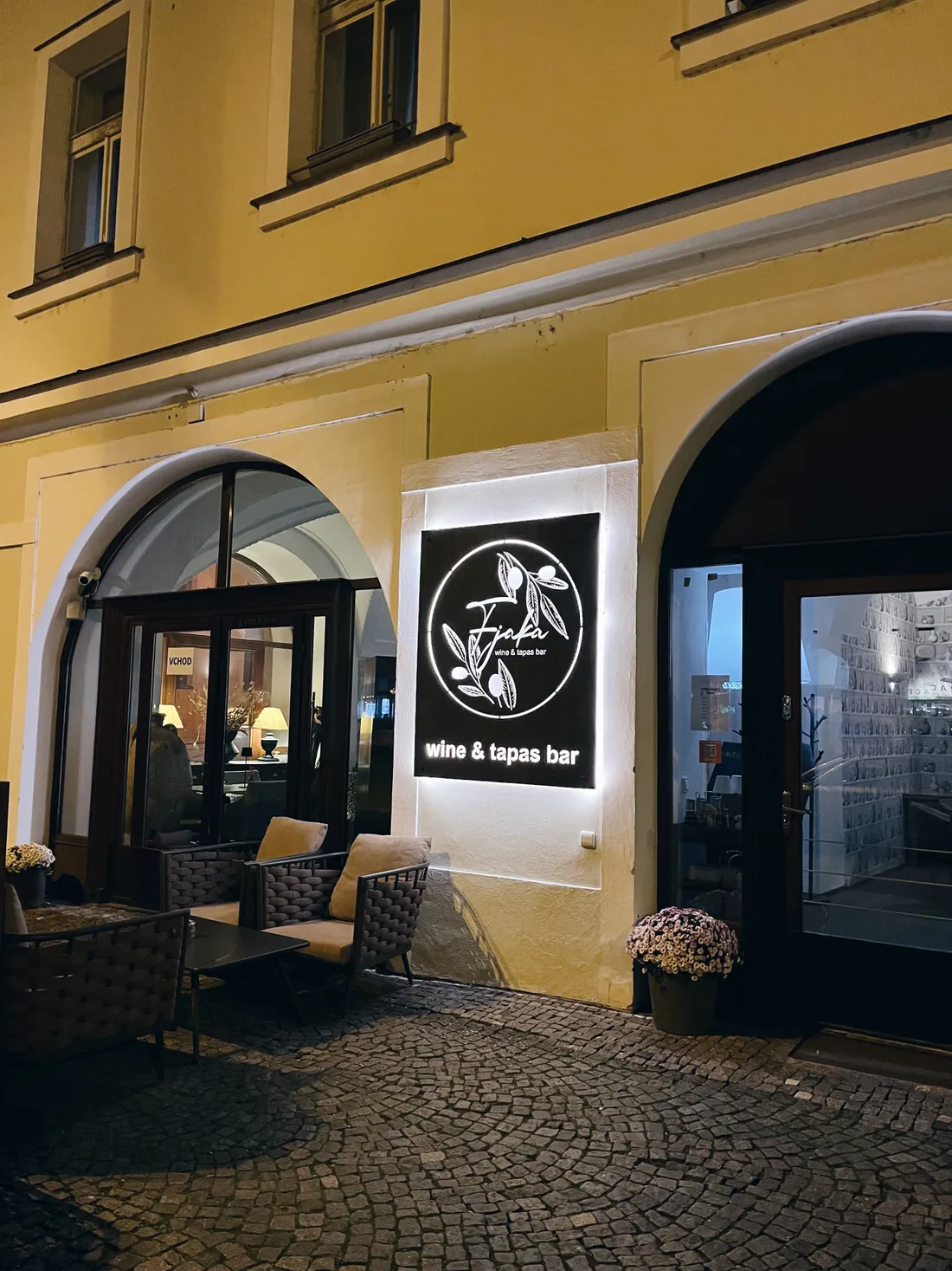 Večerní vchod do Fjaka wine & tapas bar s osvětleným logem a proutěnými křesly na chodníku