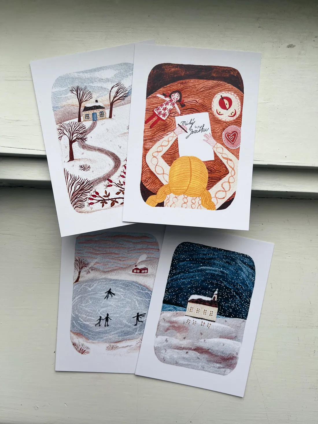 Čtyři vánoční pohlednice s ilustracemi na bílém dřevěném parapetu – chalupa, bruslení, kostel