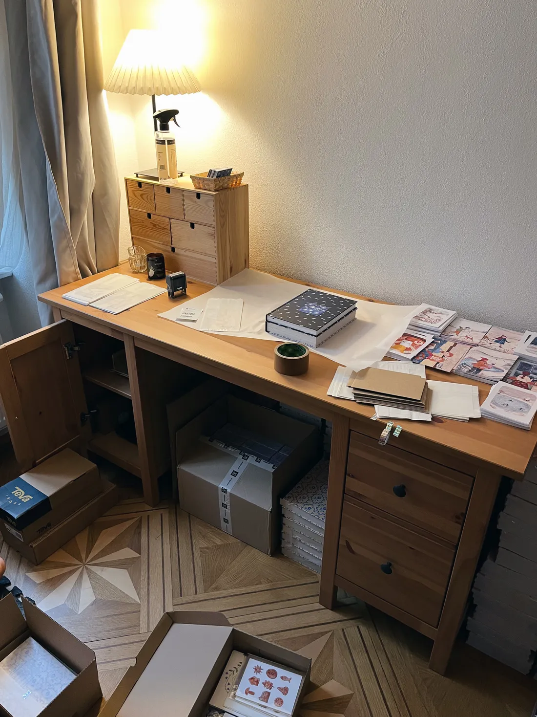 Dřevěný stůl s pohlednicemi, obálkami a balicím papírem, pod stolem kartonové krabice zásilek