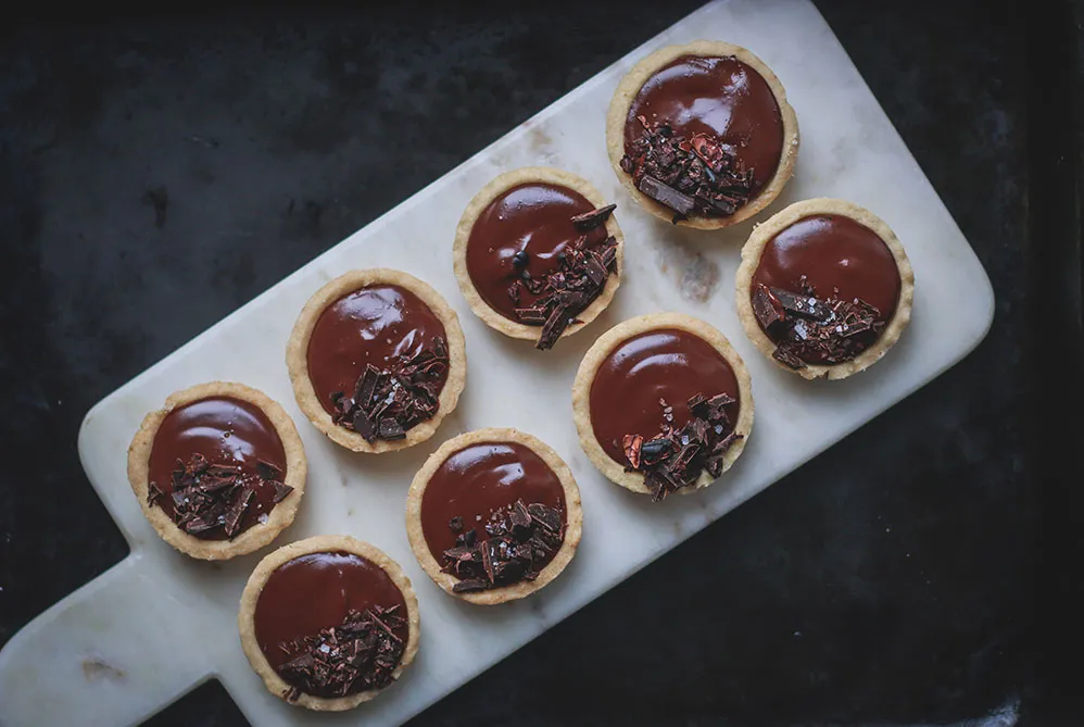 Osm mini tartaletek s ganache, kousky čokolády a mořskou solí na bílém mramorovém prkénku shora