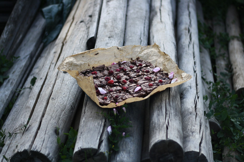 Kousky brownies s malinami a růžovými lístky na pergamenovém papíru na texturovaných šedých dřevěných kládách.