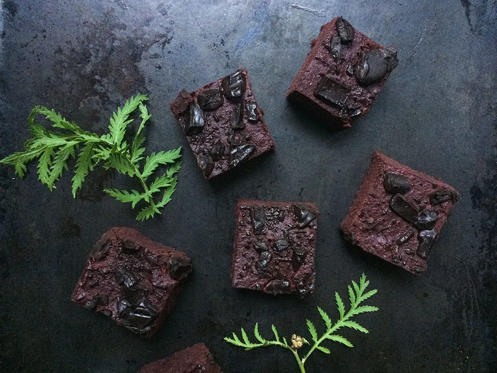 Pět čtverců brownies z červené řepy s velkými kousky čokolády na tmavém plechu a zelenými lístky bylinek.