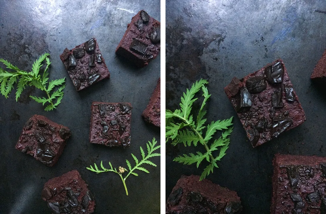 Čtverečky tmavých brownies z červené řepy s kousky čokolády na tmavém plechu se zelenými bylinkovými lístky.