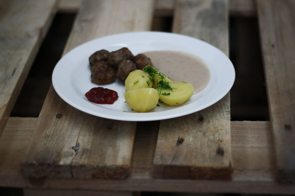 Švédské masové kuličky s bramborami, omáčkou, brusinkami a pažitkou na bílém talíři na dřevěné paletě.