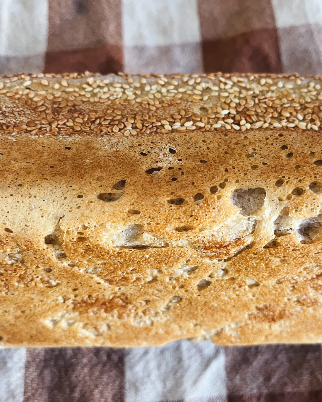 Zlatý křupavý chléb posypaný sezamem na kostkované bílohnědé utěrce, detail vzdušné kůrky a semínek
