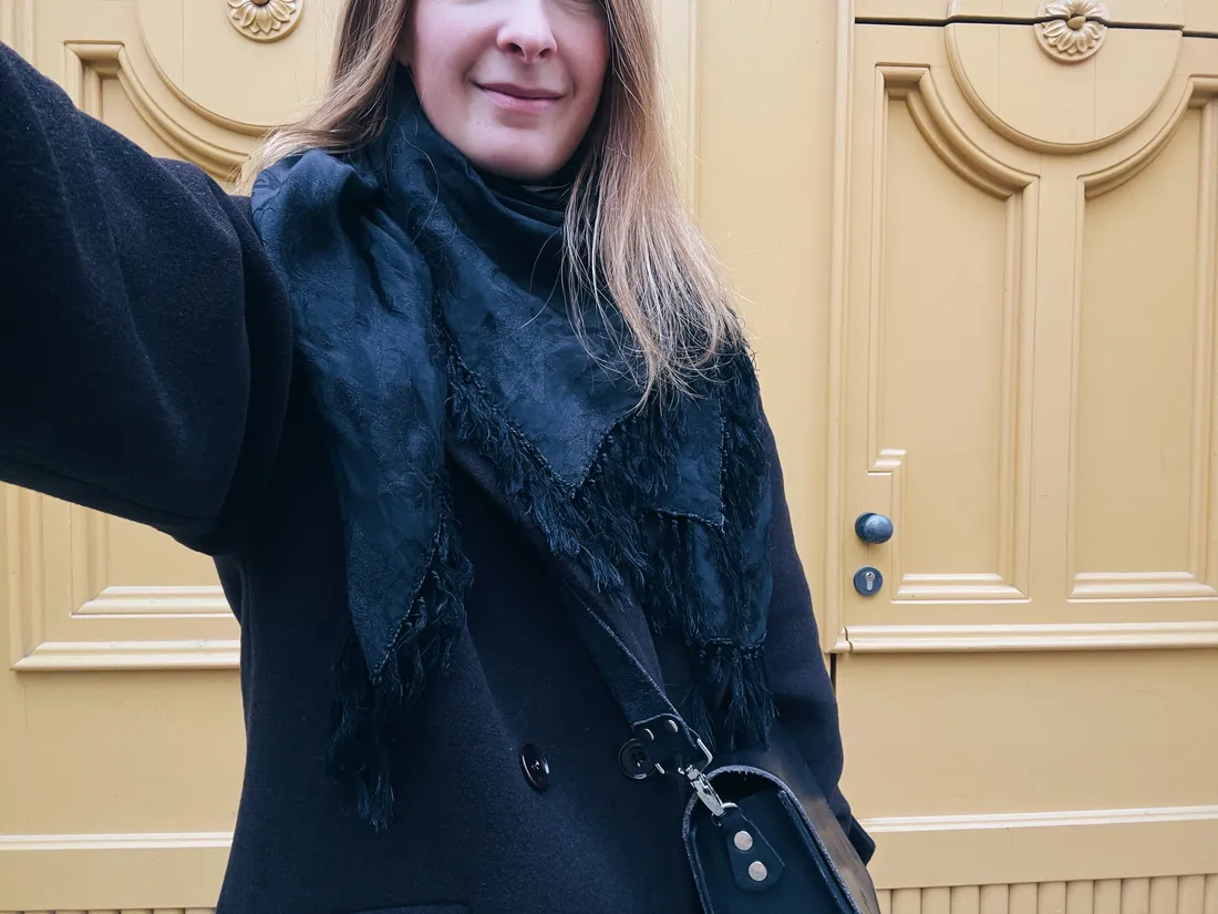 Selfie usmívající se ženy v tmavém kabátu a šálu před velkými žlutými dřevěnými dveřmi