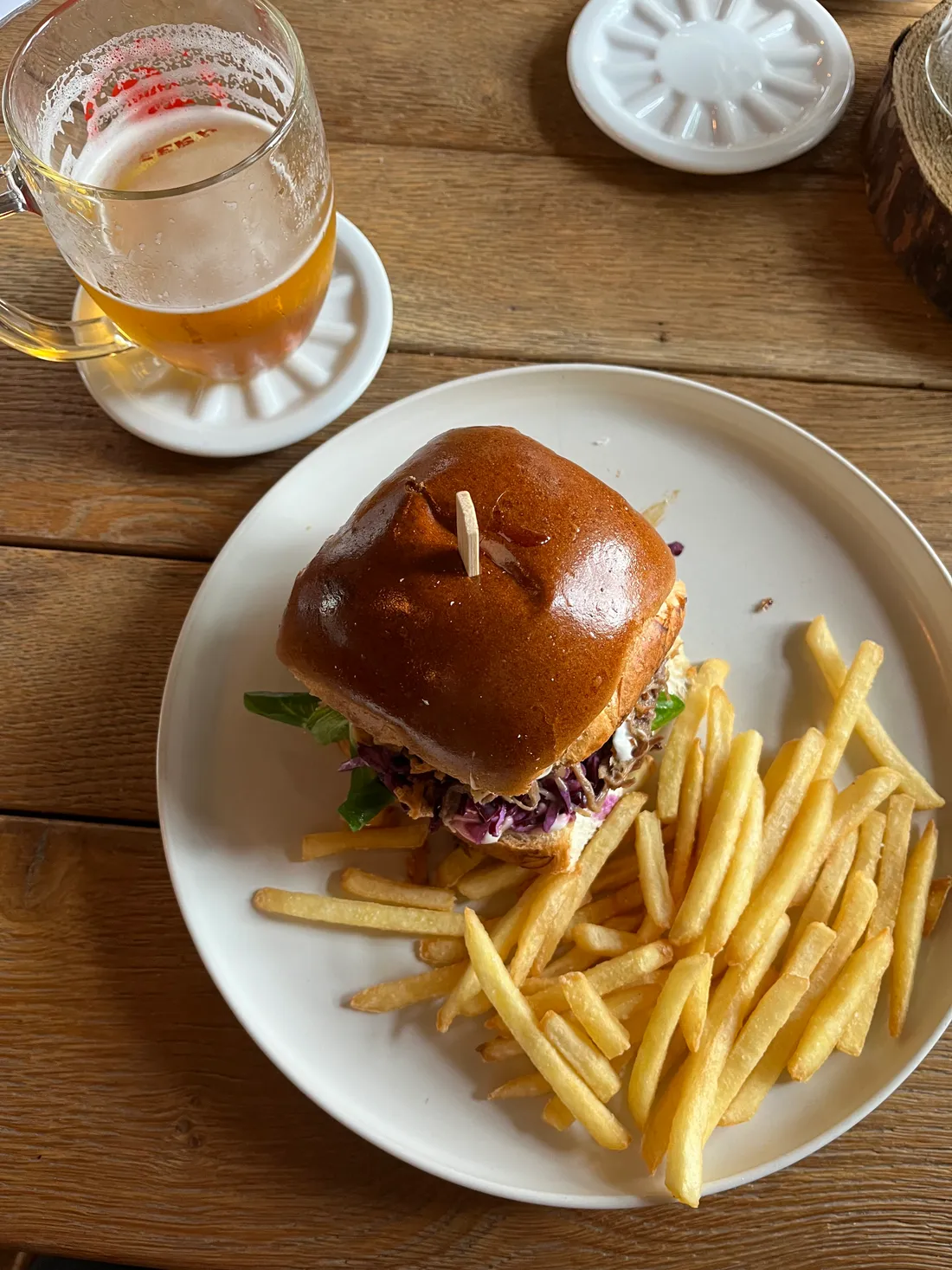 Burger s fialovým zelím a hromada hranolek na talíři, vedle sklenice piva na dřevěném stole