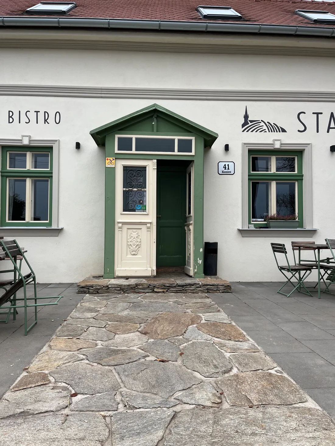 Vchod do bistra Starý kopec v Konicích s dřevěnou verandou, kamennou cestičkou a zelenými stolky