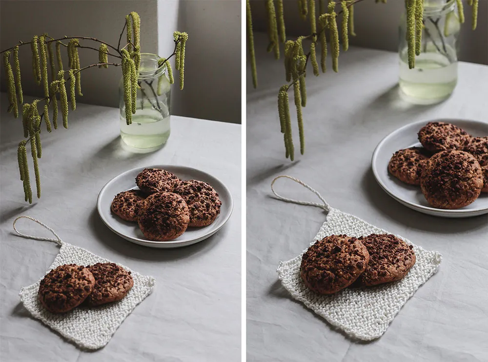 Domácí cookies s kousky čokolády na bílém talíři a pletené podložce, v pozadí sklenice s větvičkami jehněd.