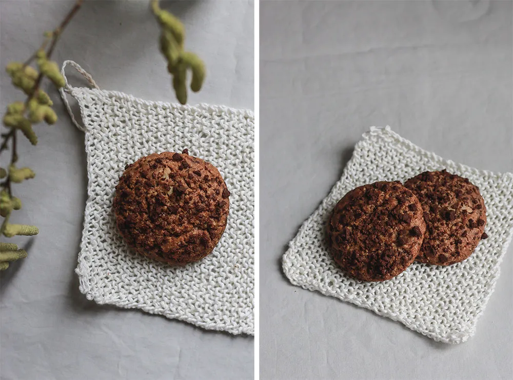 Detail dvou kulatých cookies s čokoládou na bílé pletené podložce, v těstě viditelné kousky ořechů.