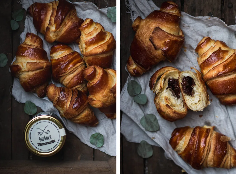 Croissanty plněné tmavou čokoládou, jeden rozlomený s náplní, vedle sklenice Šufánek na dřevěném stole s eukalyptem.
