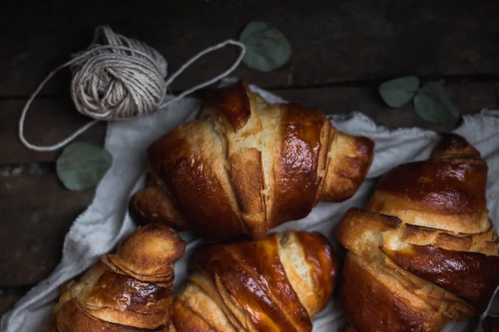 Čtyři zlatavé croissanty na lněné utěrce na tmavém dřevě, kolem provázek a listy eukalyptu v rustikální kompozici.