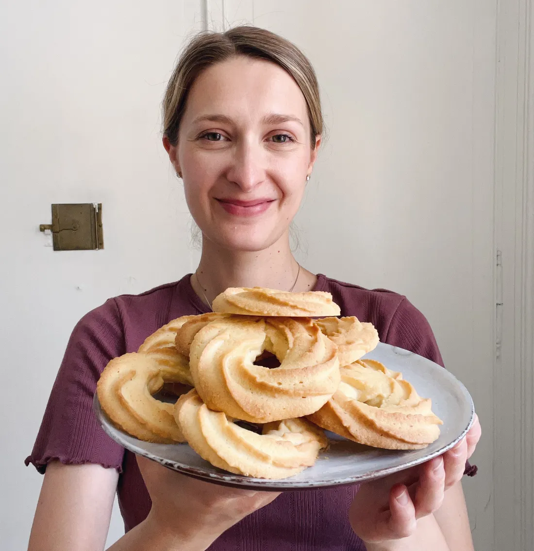 Žena v karmínovém tričku drží talíř se zlatavými dánskými vanilkovými věnečky s vroubkovaným povrchem.