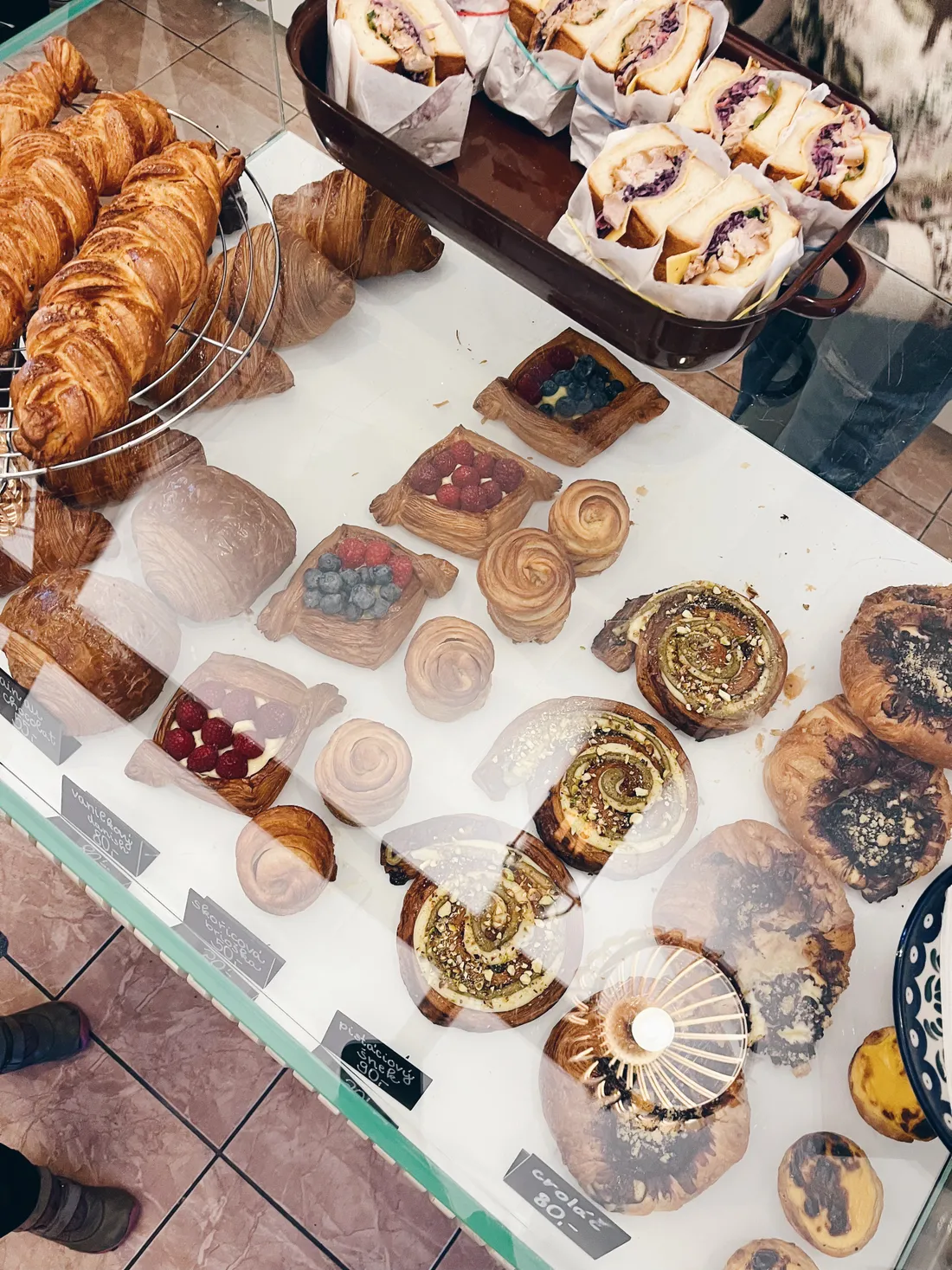 Vitrína pekárny plná croissantů, šneku s pistáciemi, ovocných tart a sendvičů s červeným zelím a cenovkami