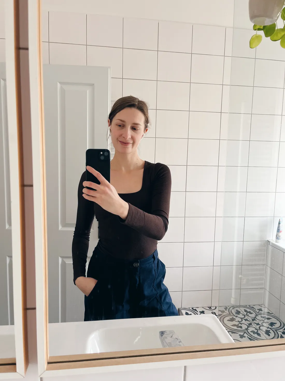 Žena v hnědém rebrovaném triku a tmavých kalhotách se fotí v zrcadle koupelny s bílými obklady a vzorovanou podlahou