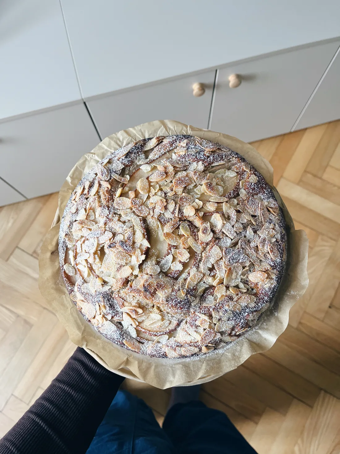 Kulatý hruškový koláč posypaný mandlovými plátky a cukrem na pečicím papíru držený v ruce nad parketami