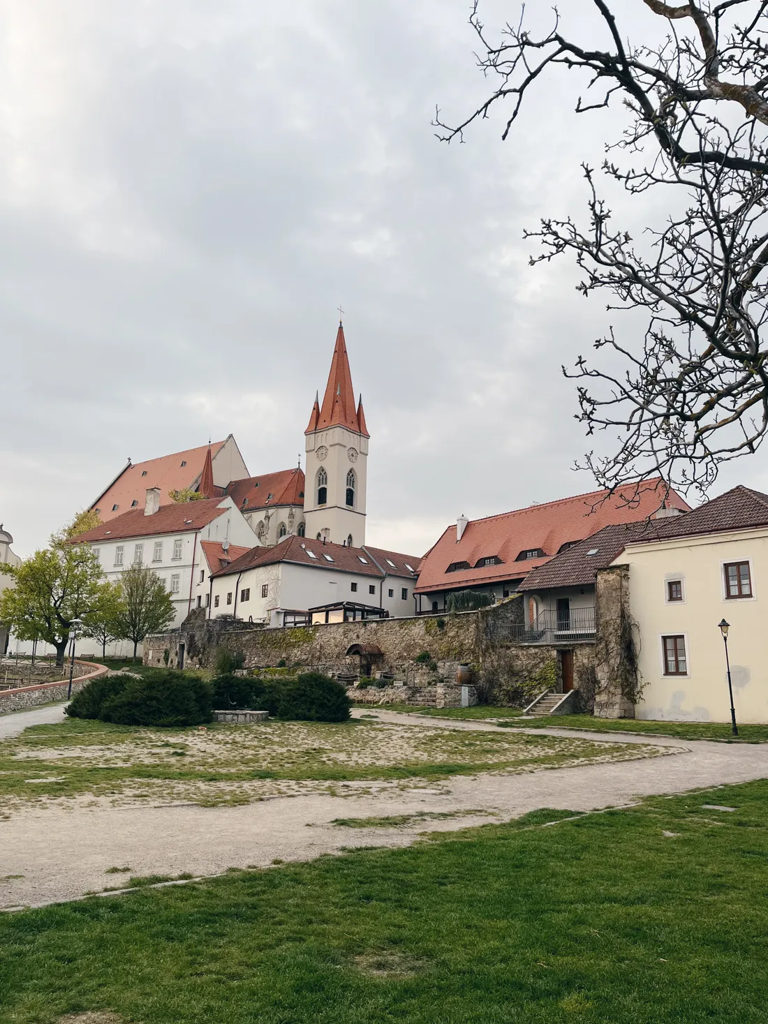 Pohled na kostel s bílou věží a červenou střechou přes kamennou zeď a parkovou cestu