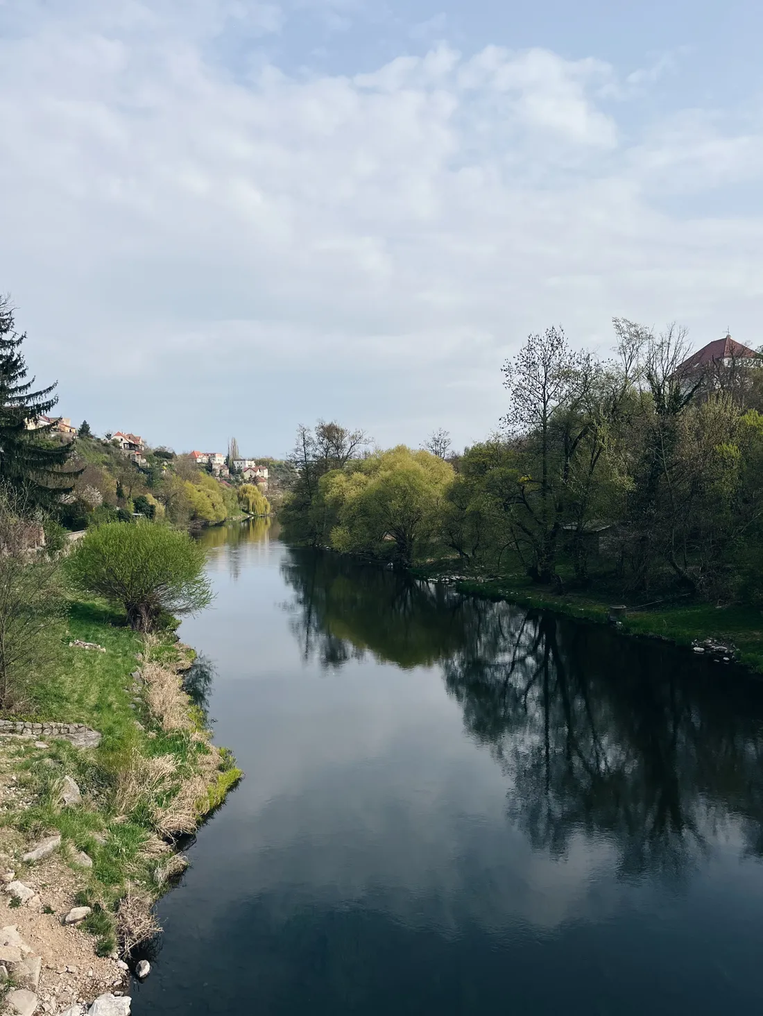 Klidná řeka odrážející stromy a oblohu, na březích zelené keře a domy ve svahu