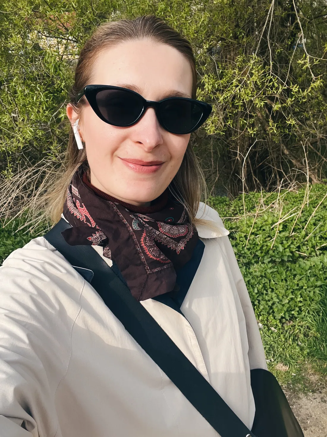 Selfie ženy v béžovém trenčkotu, slunečních brýlích a hnědém šátku mezi jarními keři