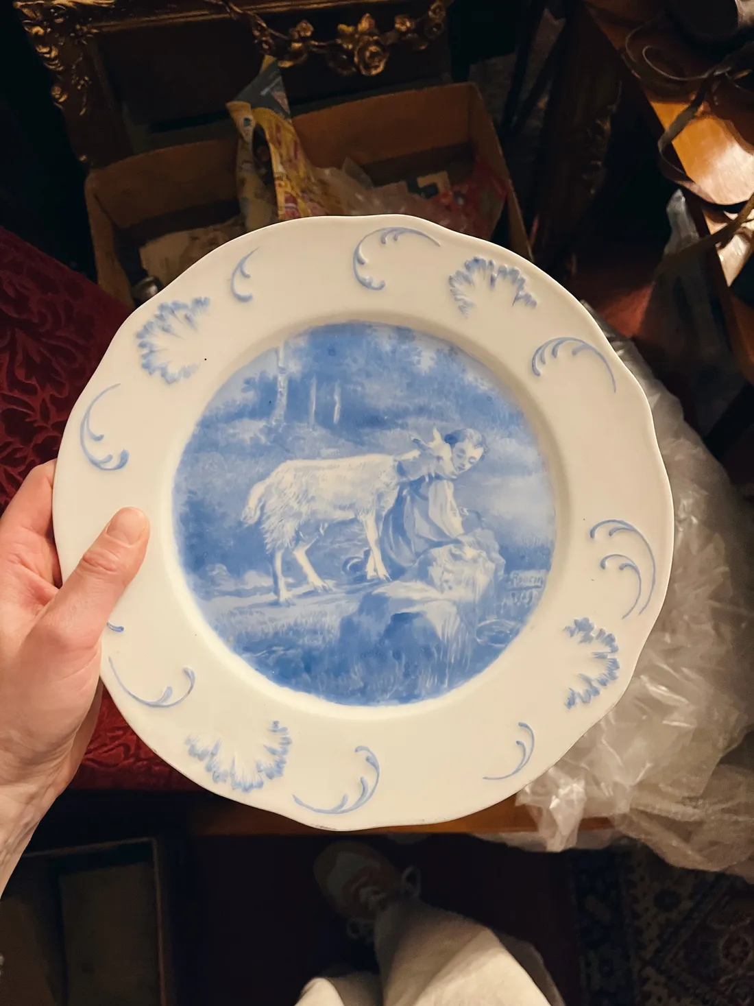 Ruka drží bílý porcelánový talíř s modrým pastoralním motivem dívky a kozy na okraji