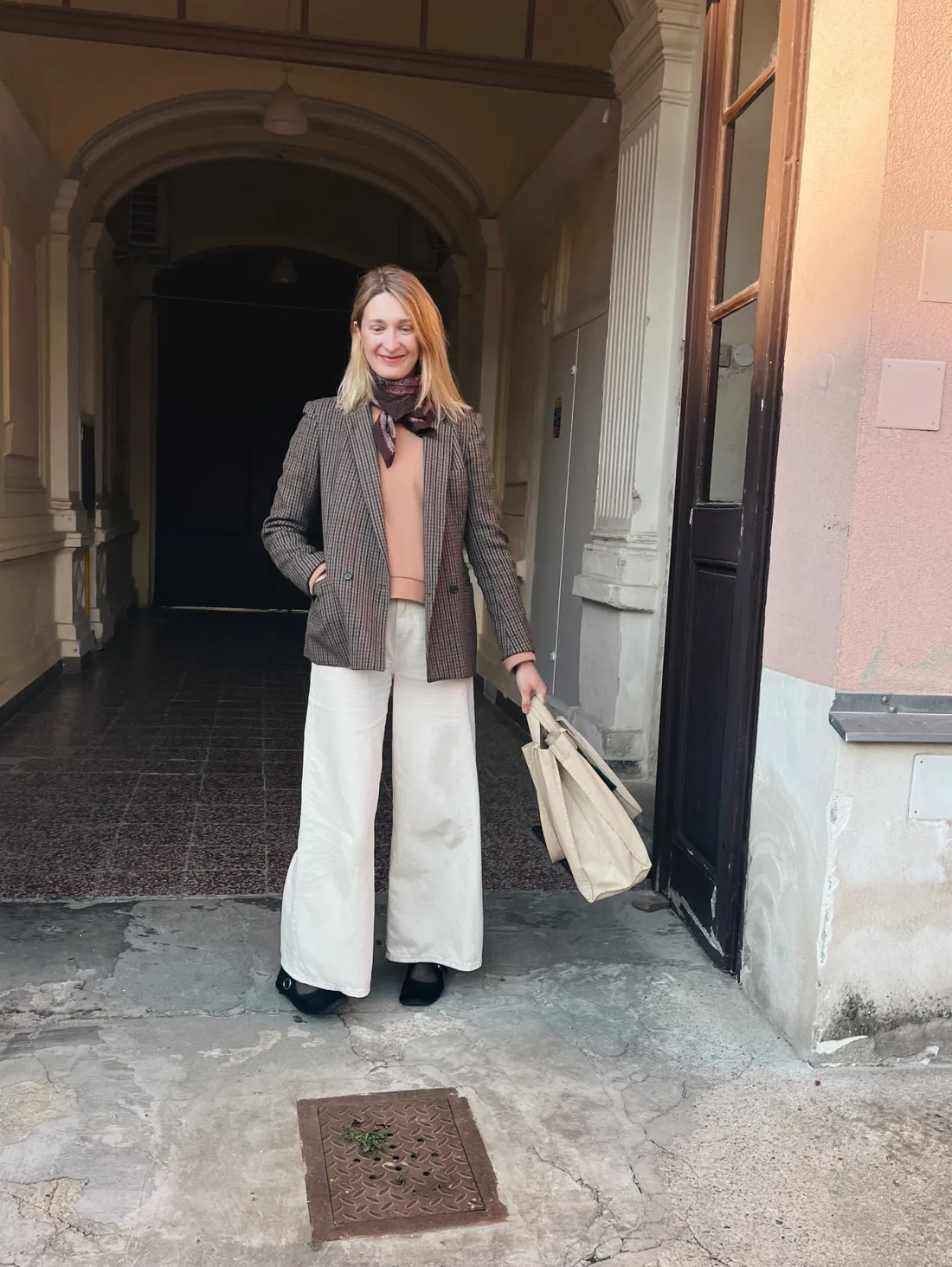 Žena v hnědém blazeru a krémových kalhotách s taškou v klenutém průchodu historické budovy
