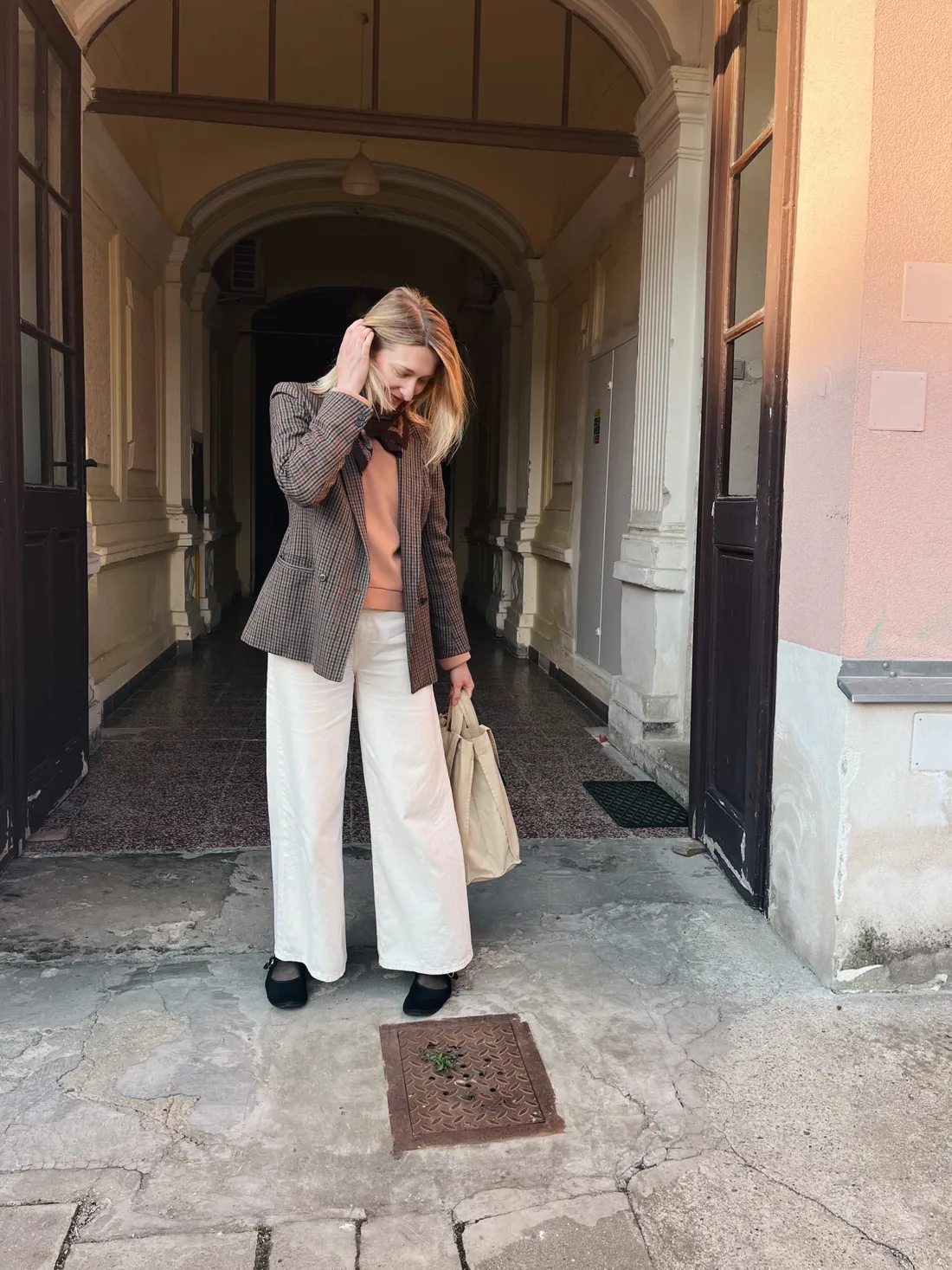Blond žena v kostkovaném blazeru a bílých kalhotách upravuje vlasy v klenutém průchodu