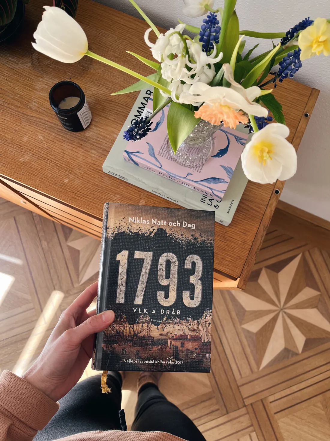 Ruka drží knihu 1793 nad dřevěným stolkem s kyticí tulipánů a modřenců na parketové podlaze