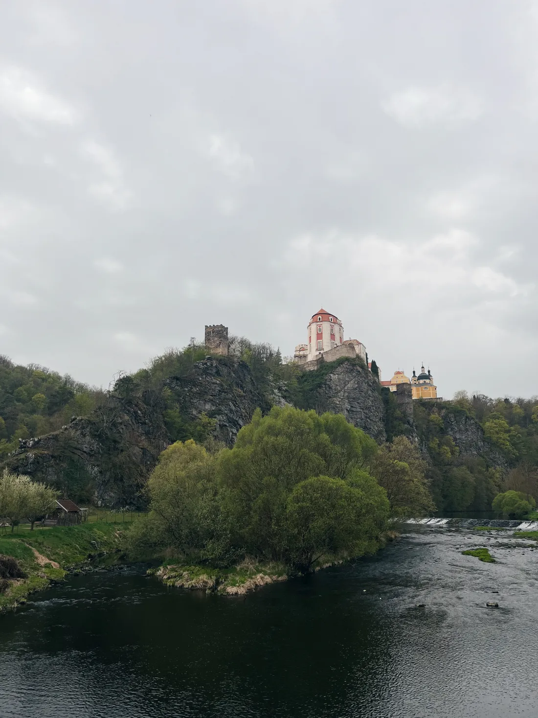 Zámek ve Vranově nad Dyjí na skalním útesu nad řekou s weirem a zeleným ostrovem pod šedou oblohou