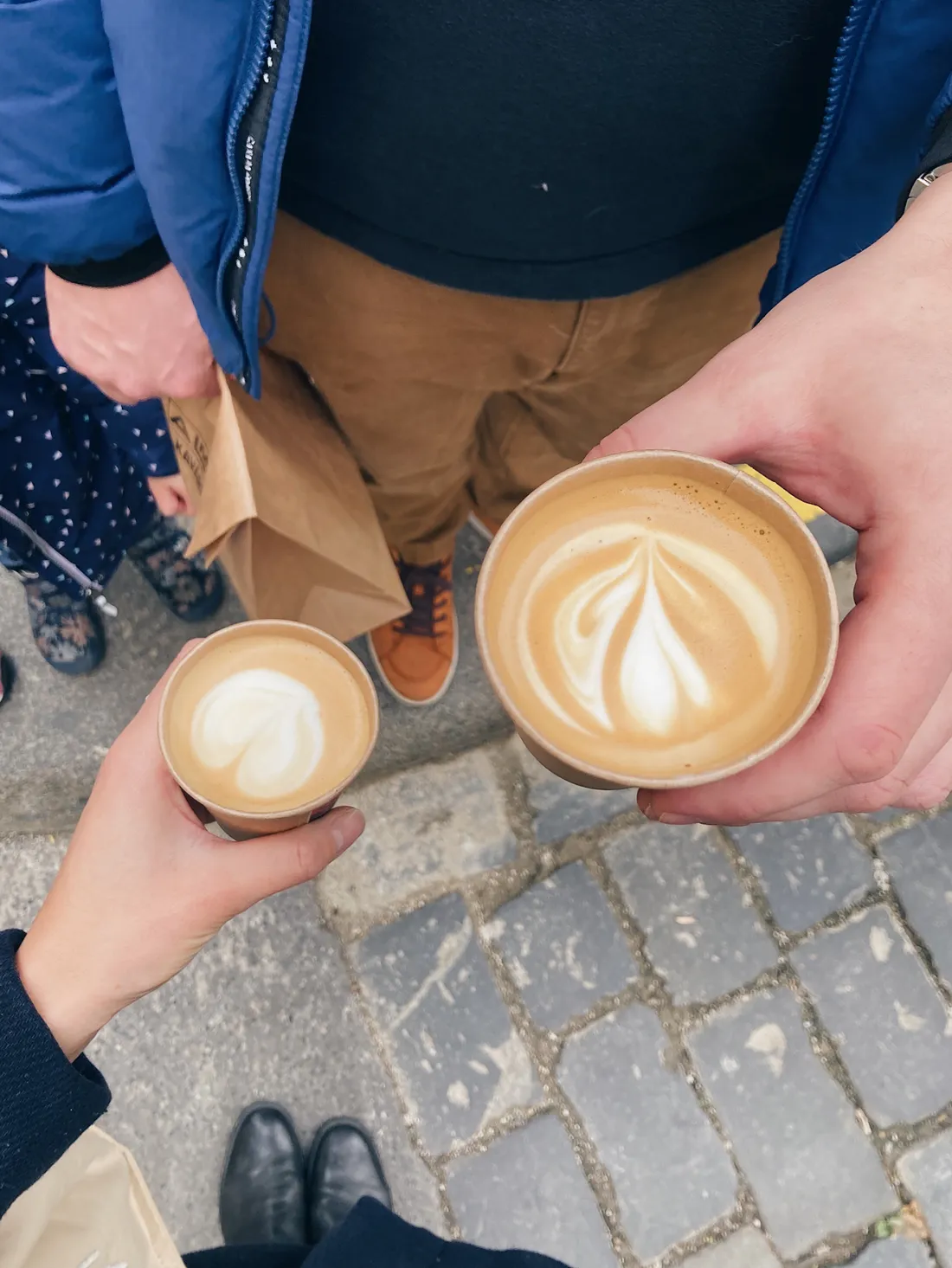 Dvě papírové kelímky cappuccina s latte art v rukou nad dlážděným chodníkem, vedle hnědé tašky a dítěte