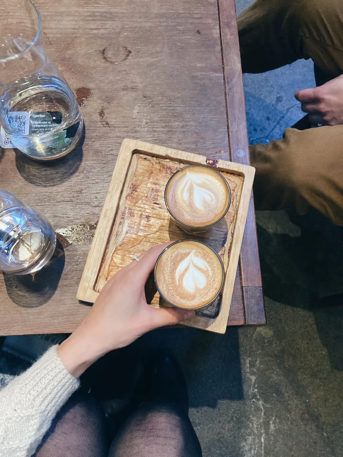 Dvě sklenice cappuccina s latte art ve tvaru srdce na dřevěném podnosu, ruka v bílém svetru sahá po šálku