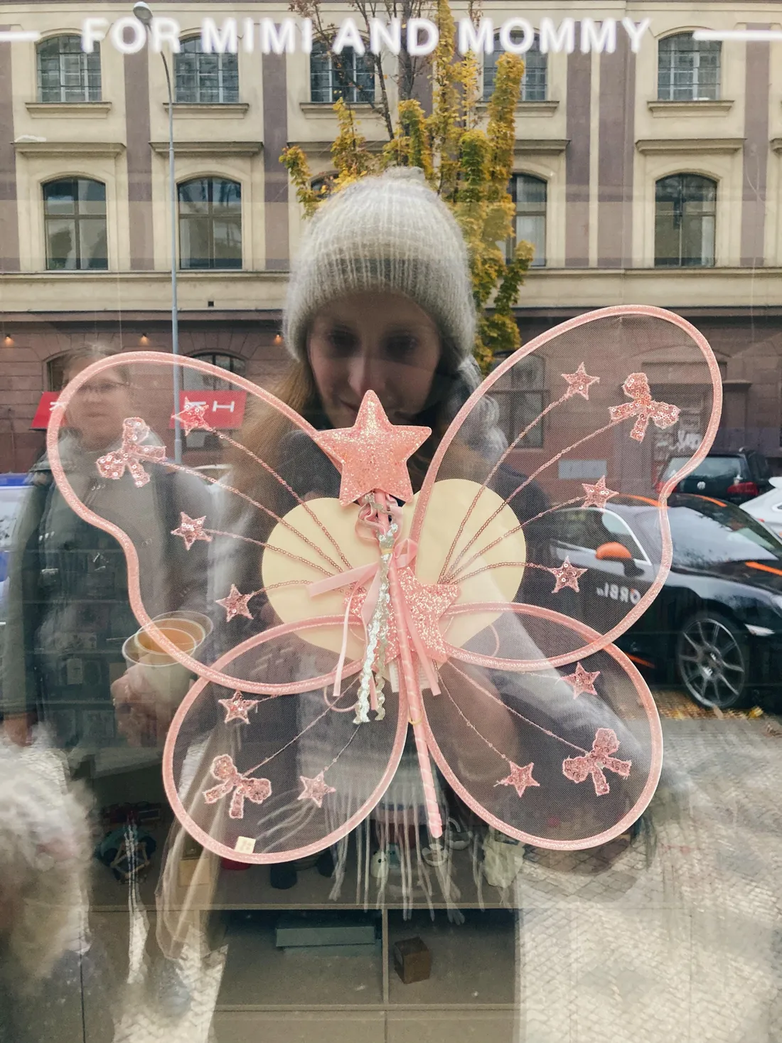 Růžová vílí křídla a hůlka na výloze obchodu For Mimi and Mommy, za sklem žena v čepici a podzimní ulice