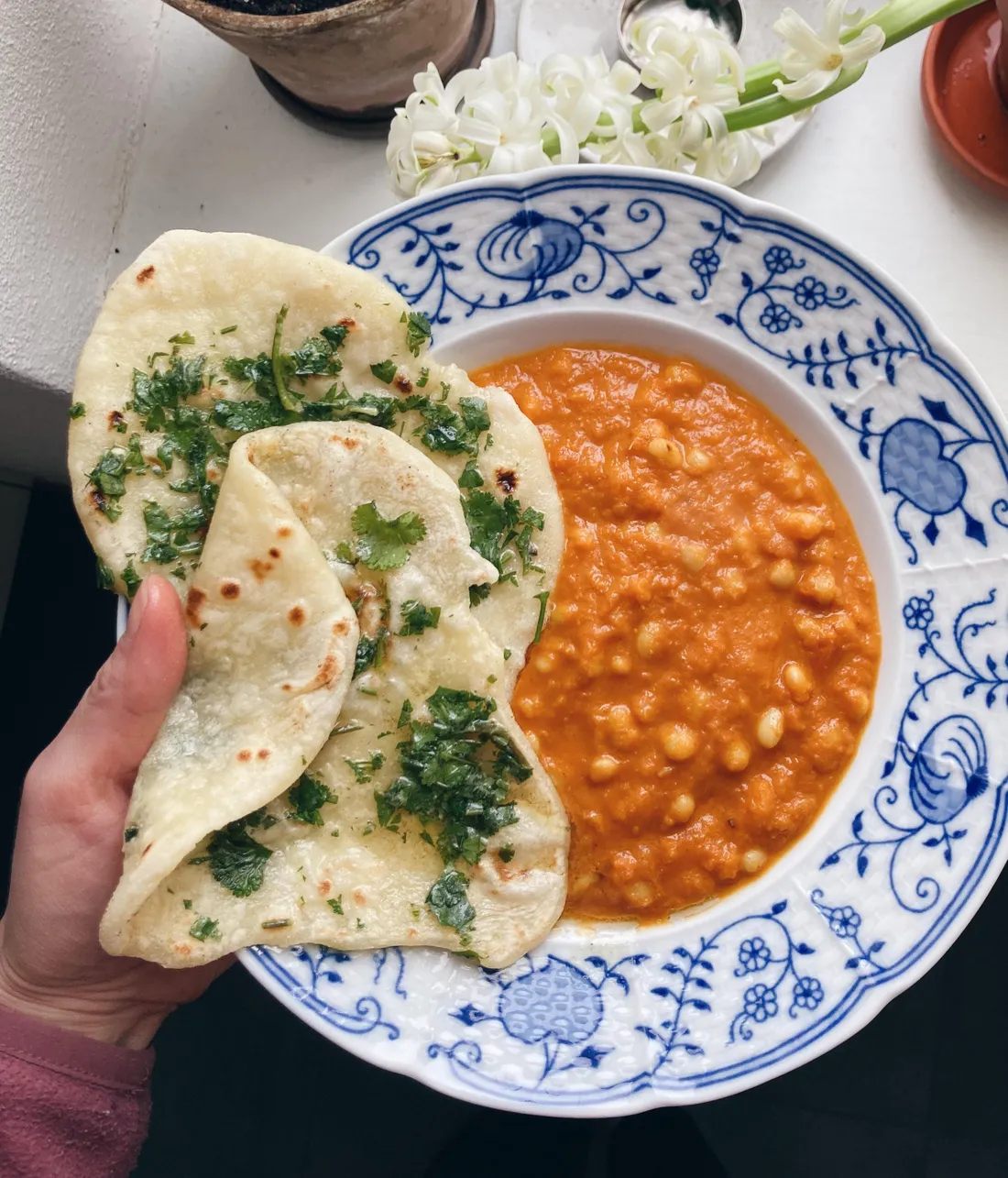 Ruka drží dva kousky naanu s bylinkami nad modrobílým talířem s oranžovou fazolovou omáčkou a bílými květy v pozadí