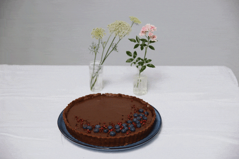 Čokoládový tart s borůvkami a růžovým pepřem na modrém talíři, vázičky s květy za ním