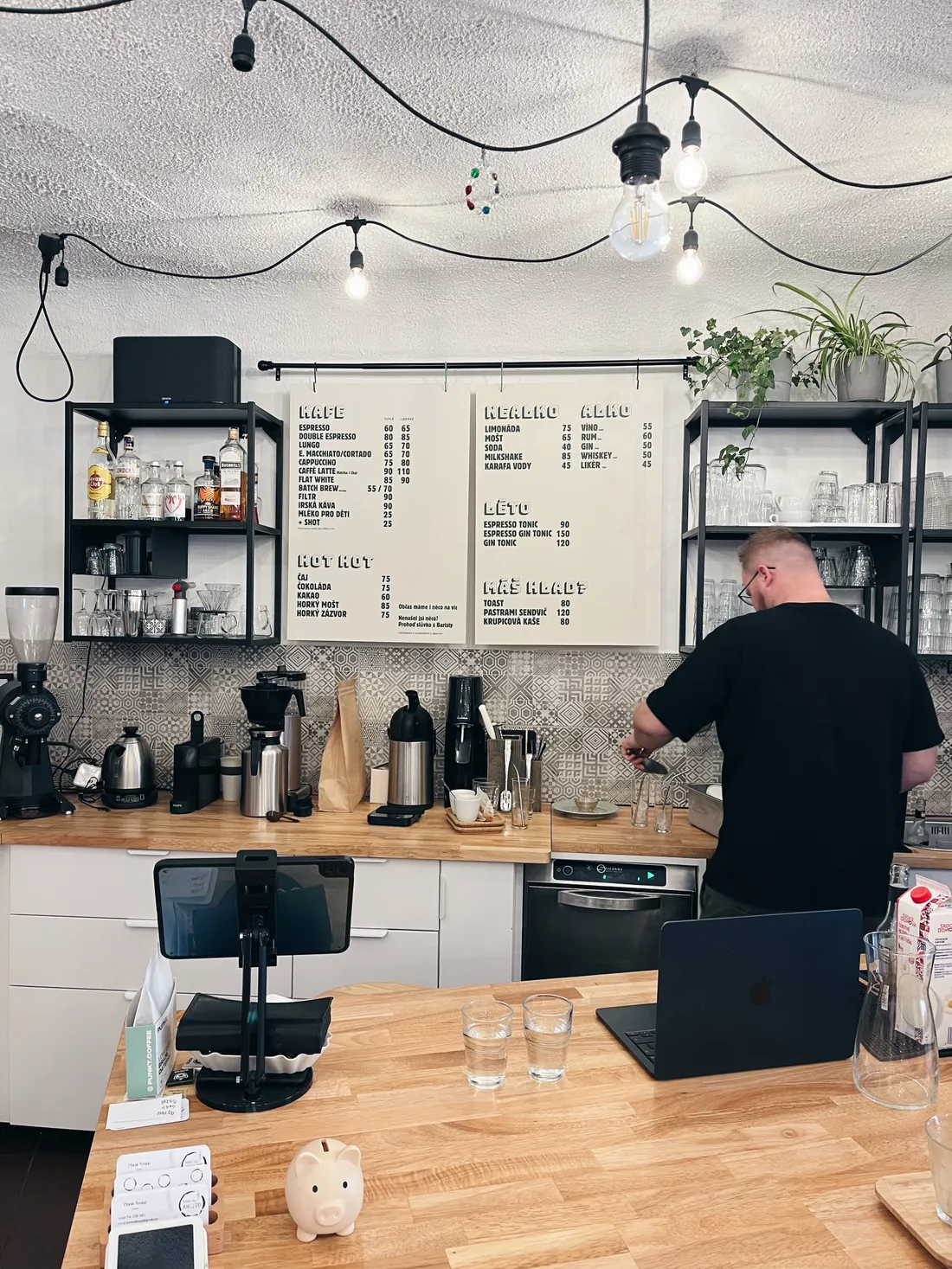 Interiér kavárny s baristou za dřevěným pultem, menu na tabuli a řetězovými žárovkami