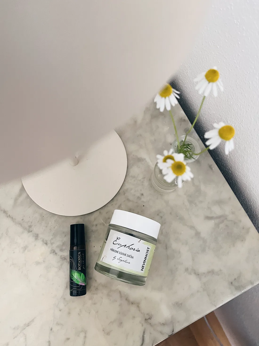 Sójová svíčka Soaphoria Nevinnost a roll-on Botanica na mramorovém stolku, vázička heřmánku